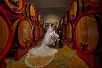 recently married couple siting in a wine celar surrounded by huge wooden wine barrels